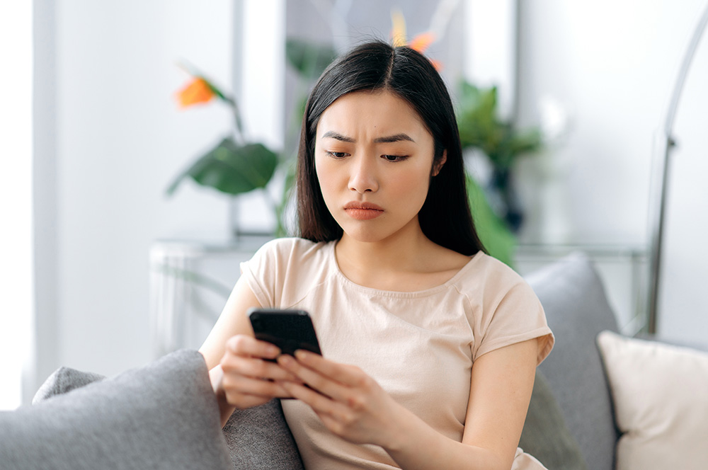 Young woman sits on couch with phone in hand debating whether to call the emergency room or an urgent care clinic.